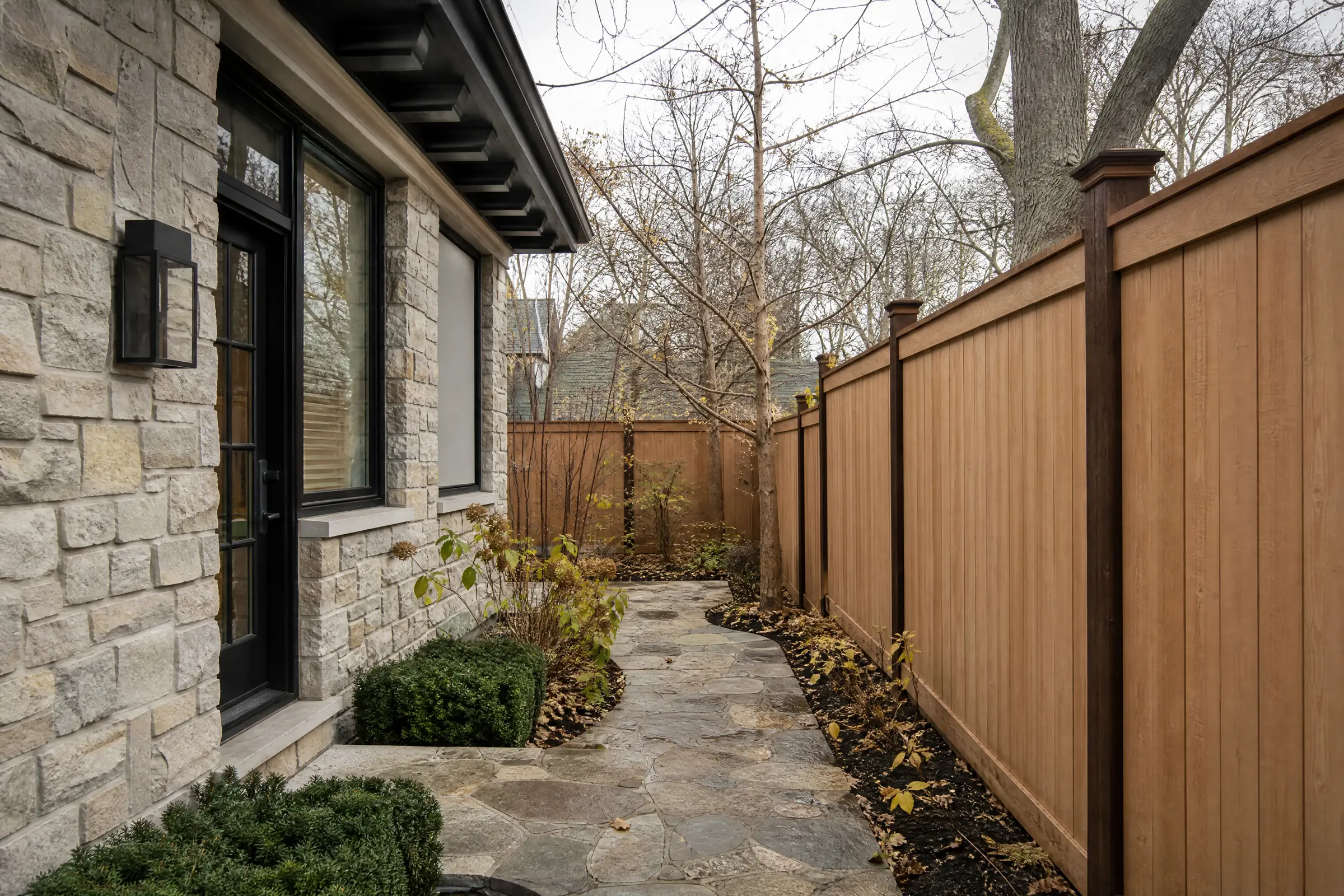 Cedar privacy fence freshly stained beside a fieldstone house in late autumn